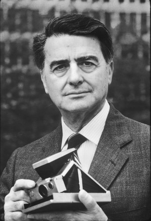 A close perspective black and white photo of a man wearing a suit and tie looking thoughtfully at the viewer, a vintage polaroid camera in his hands.