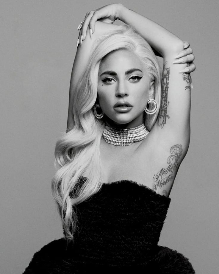 A black and white photo of artist Lady Gaga in a black strapless dress and long curled blonde wig draped over her right shoulder. Her arms are over her head and she is looking directly at the camera.