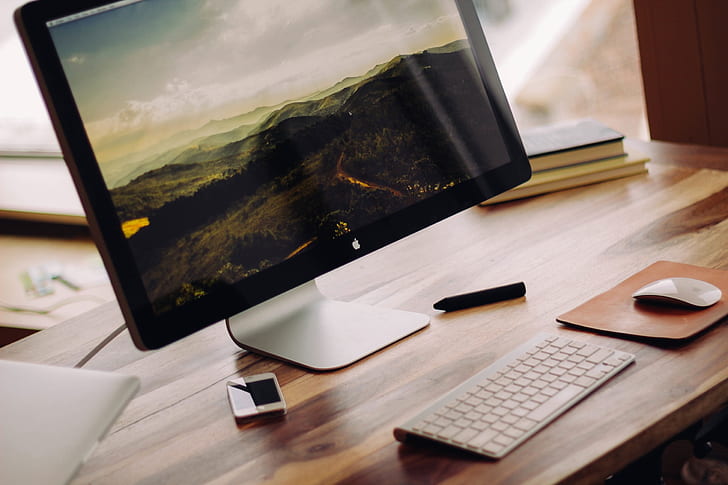 A black and white photo of an iMac on a desk with a keyboard and mouse on a mousepad. There is a black crayon laying in front of the mousepad.
