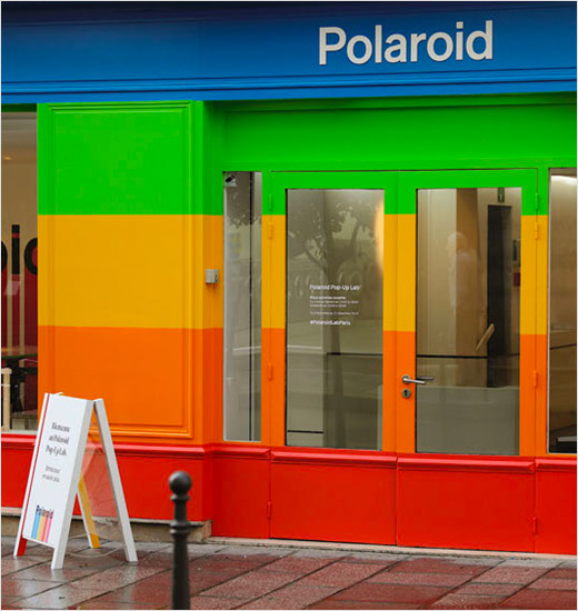 A storefront that has bright blue, green, yellow, orange, and red horizontal stripes from sidewalk to over the door on the outside of the building. The Polaroid text logo appears over the door with a sign out front on the sidewalk.